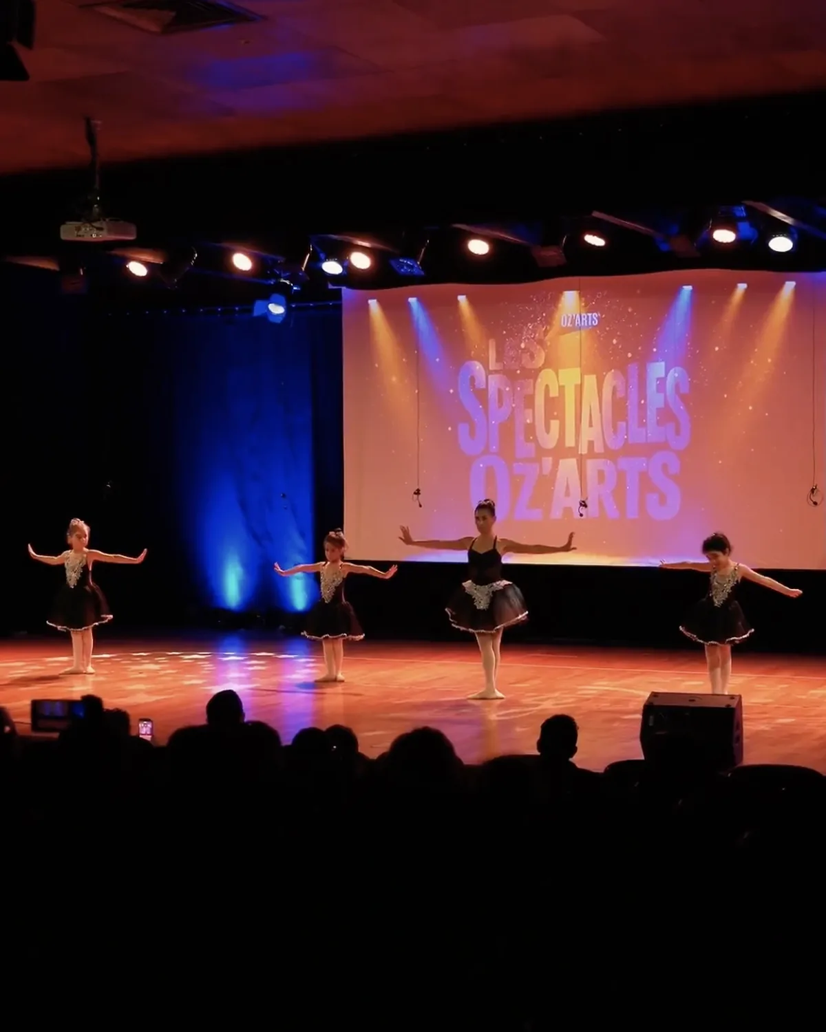 Quatre jeunes danseuses en tutu noir se produisent sur scène devant le logo des Spectacles OZ'ARTS.